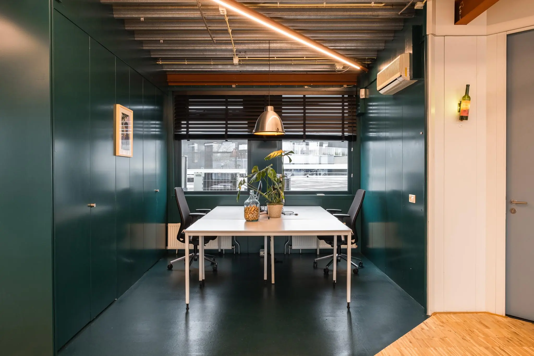 Vergaderruimte aan de Johan van Hasseltweg met een strak wit bureau, twee bureaustoelen, groene muren en een grote hanglamp boven de tafel.